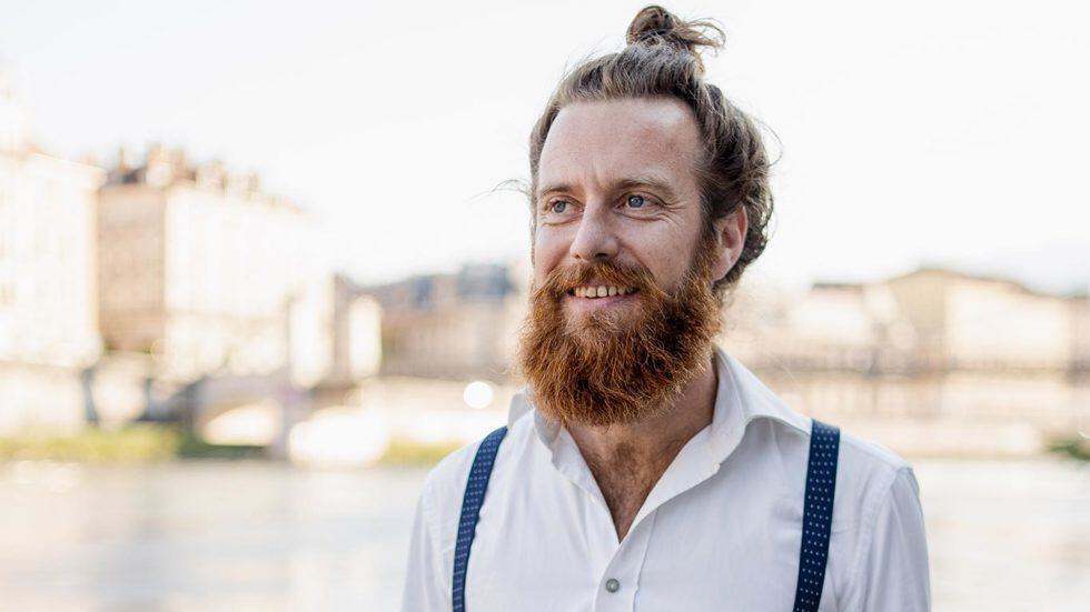 Homme souriant avec barbe, en tenue décontractée, dans un environnement urbain au bord de l'eau.
