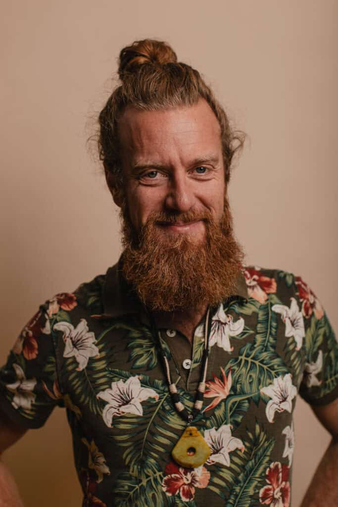 Homme avec barbe et cheveux en chignon, portant un t-shirt à motifs floraux et un collier en bois.