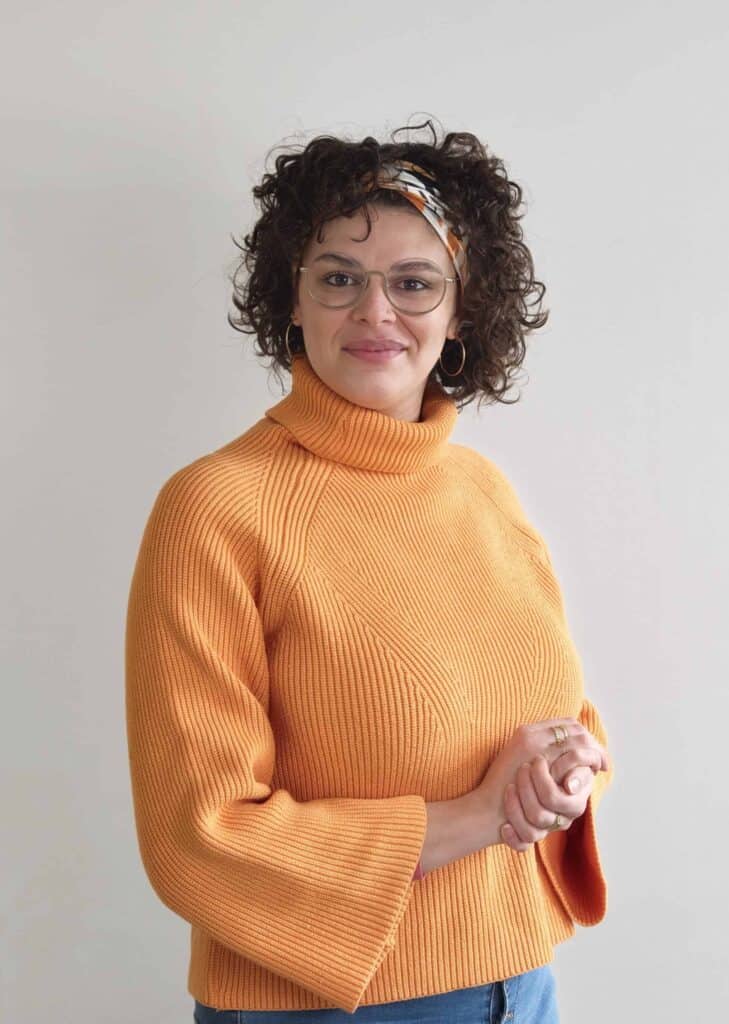 Femme avec lunettes et foulard, portant un pull orange, dans un cadre neutre.