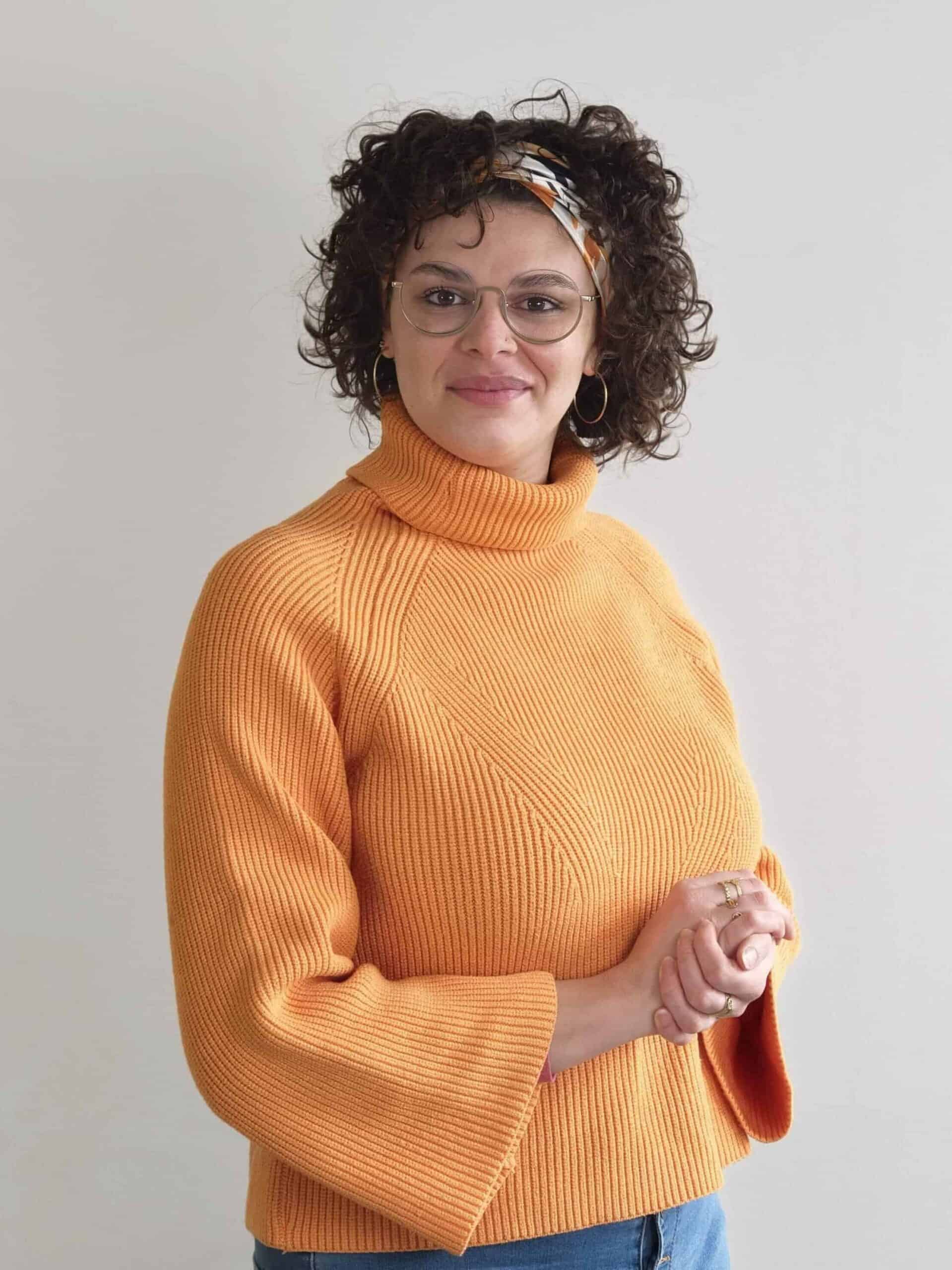Femme souriante portant un pull orange et des lunettes, posant avec confiance.